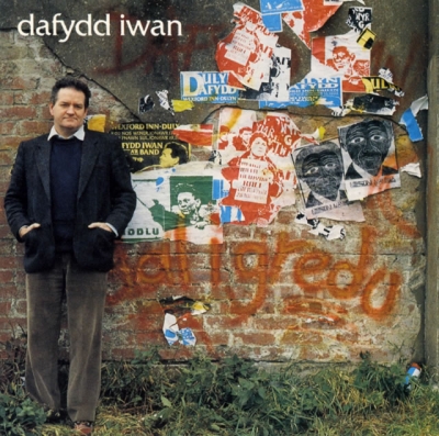 A man in a dark coat stands beside a weathered wall layered with torn posters, graffiti and painted marks.